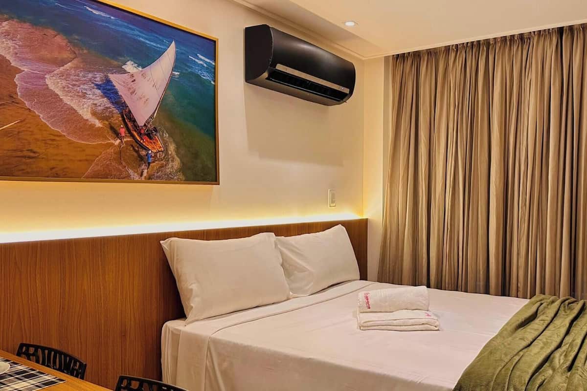 Una habitación de hotel con una cama, un cuadro de un velero, un aire acondicionado y una cortina.