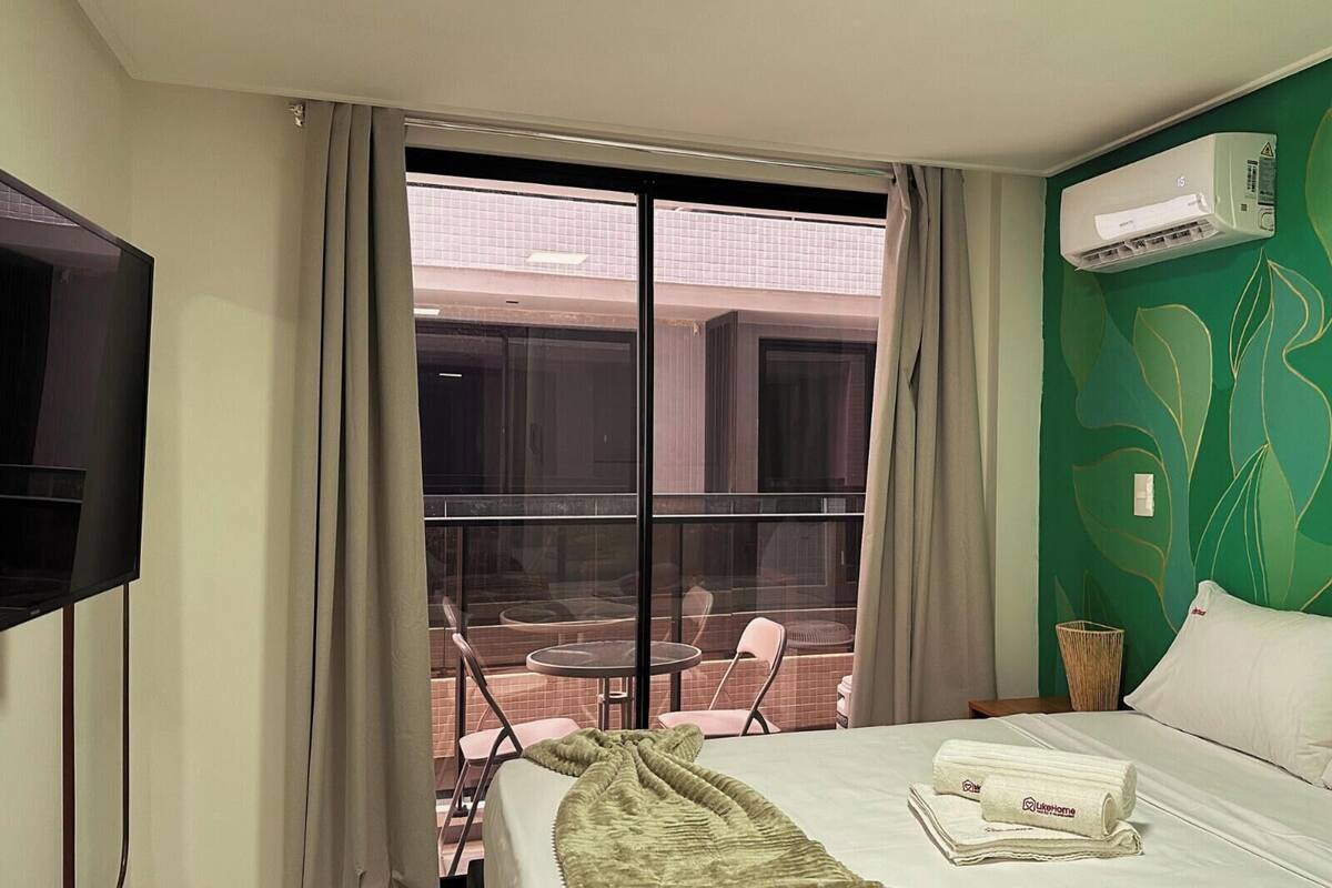 Una habitación de hotel con una cama, un televisor, una ventana con cortinas, una pared verde y un equipo de aire acondicionado.