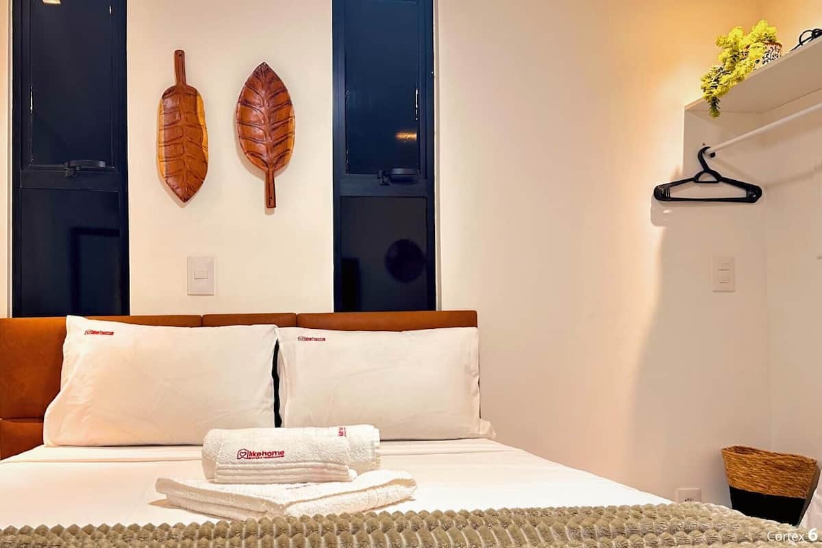 Una cama bien hecha con ropa blanca y una toalla doblada, dos adornos de madera con forma de hojas, un perchero negro y una pequeña cesta a la derecha.