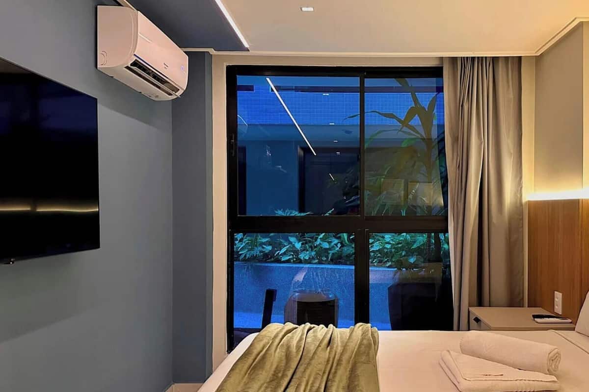 Una habitación de hotel con cama, televisión, ventana con cortinas y aire acondicionado instalado en la pared.