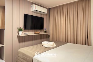A hotel room with a bed, a mounted TV, an air conditioning unit, a shelf with a plant and a vase, and a curtain. - Hit Cabo Branco III - LikeHome (João Pessoa)