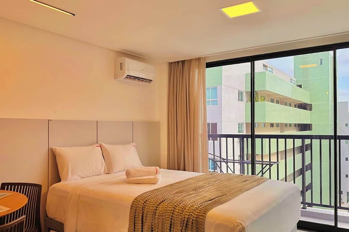 Habitación de hotel con cama, una mesa con platos y vasos, unidad de aire acondicionado y vista a un edificio a través de un ventanal grande.