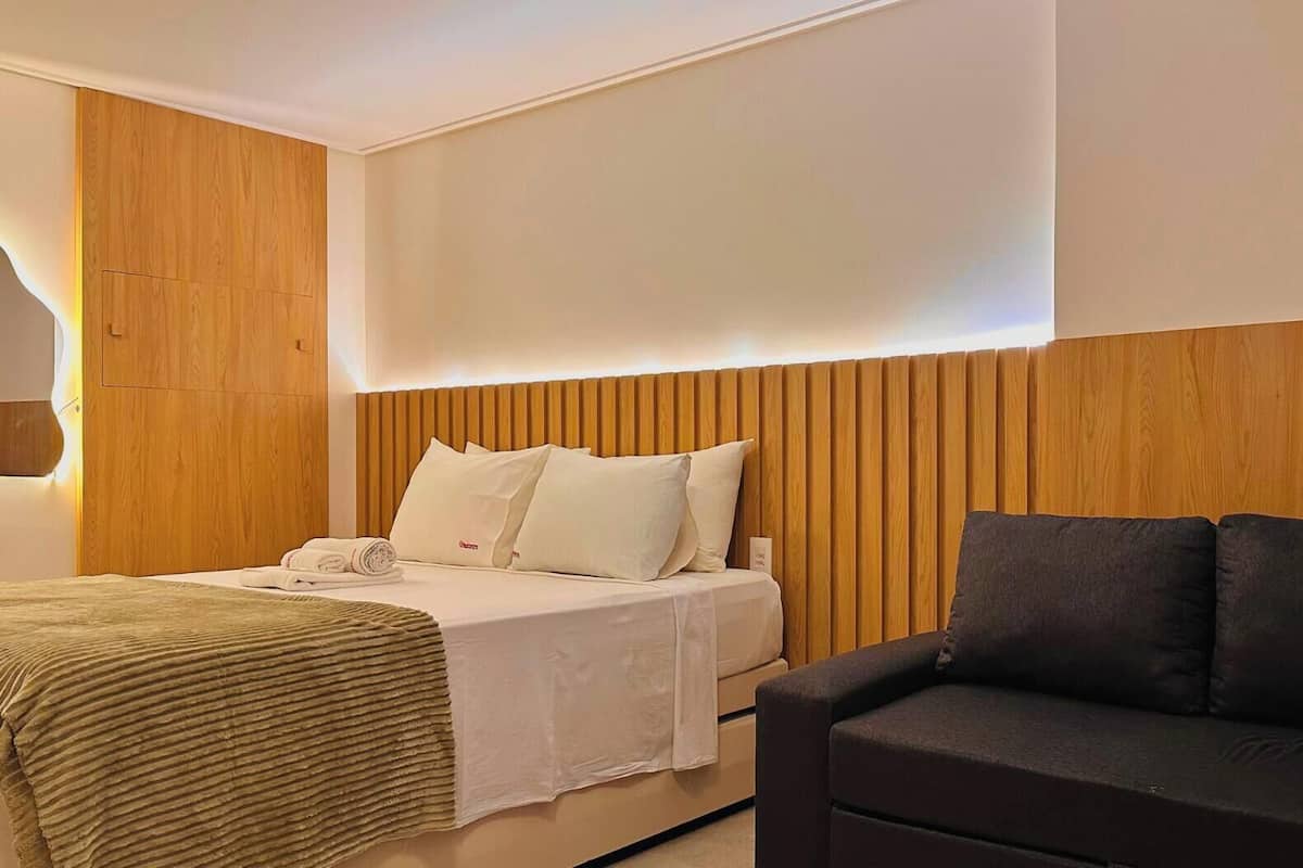 Una habitación de hotel con una cama, un sofá negro, paneles de madera y un cabecero blanco.