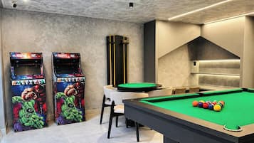 Mesa de bilhar com bolas, duas máquinas de jogos eletrônicos, mesa de sinuca e uma área de estar.