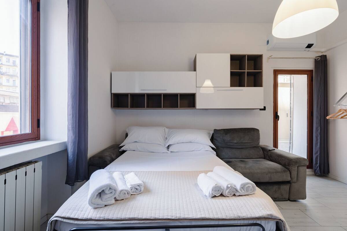 Une chambre d’hôtel avec un lit, un fauteuil gris, une armoire et un luminaire suspendu.