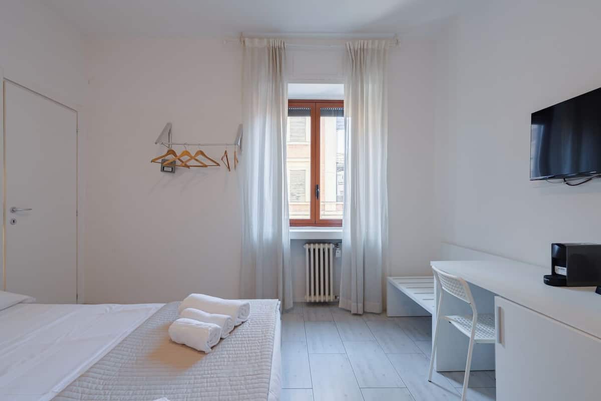 Une chambre d’hôtel avec un lit, un bureau, une chaise, un radiateur et une fenêtre avec des rideaux.