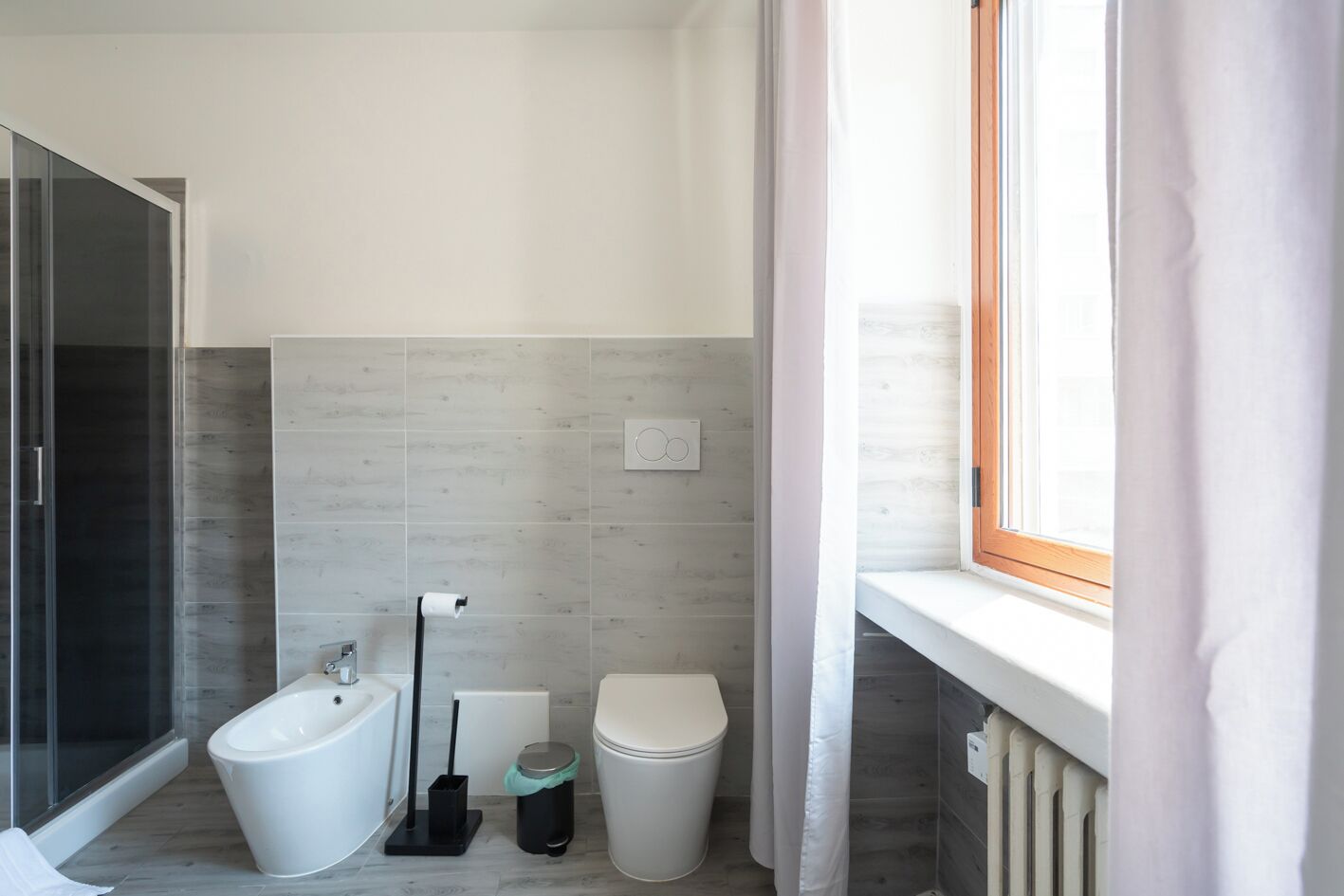 A modern bathroom with a shower, toilet, bidet, and a window with curtains.