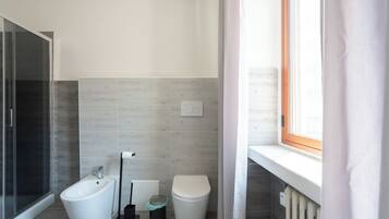 A modern bathroom with a shower, toilet, bidet, and a window with curtains.