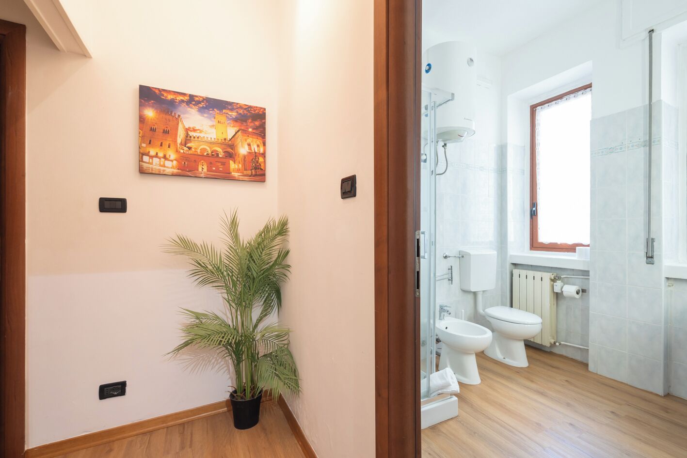 A hallway with a potted plant, a painting, and a bathroom with two toilets and a shower.