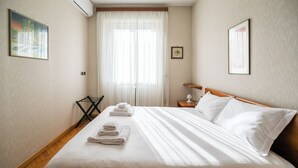 Ett hotellrum med en snyggt bäddad säng, ett fönster med tunna gardiner, en väggmonterad luftkonditioneringsanläggning och ett litet bord med en lampa.