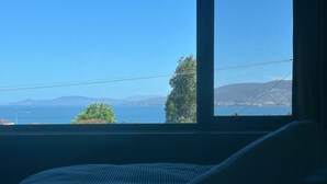 A room with a large window overlooking a coastal landscape, including a body of water and distant hills. - The Blue Window - 15 Mins to CBD&Airport (Howrah)