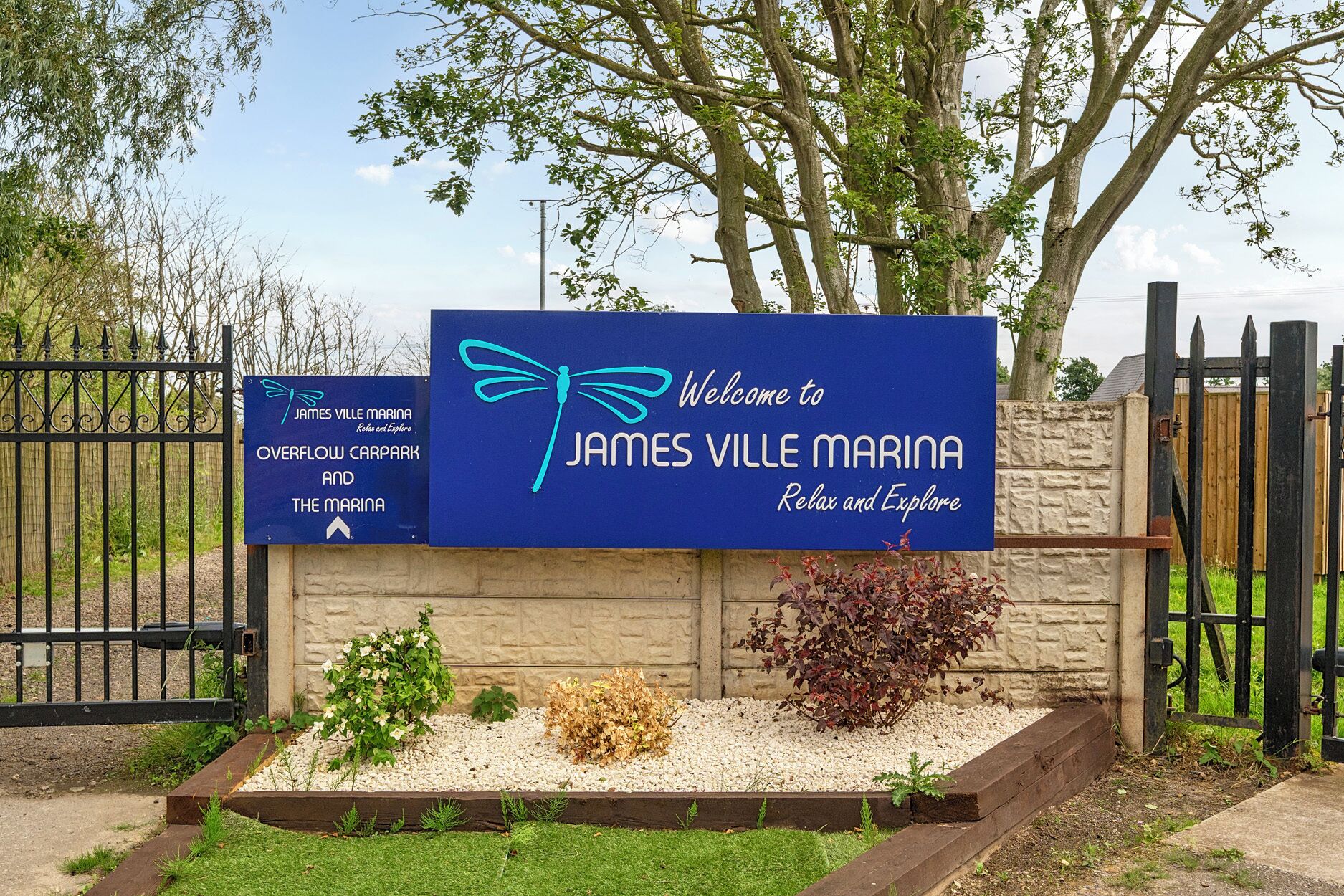Une signalisation accueillante pour la marina de James Ville, avec un logo représentant une libellule, ainsi qu’une signalisation pour le parking annexe et la marina, et une entrée fermée par un portail, avec un mur de pierre et de la verdure.
