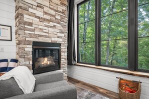 A stone fireplace with a fire, a large window with a forest view, and a gray sofa.