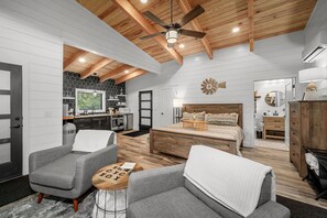 Une chambre spacieuse avec un grand lit, deux fauteuils gris, une table basse en bois et un ventilateur de plafond.