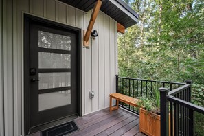 A wooden deck with a bench and a potted plant, a black door with a window, and a railing.