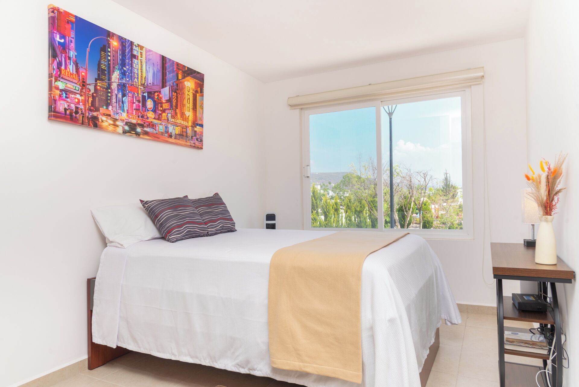 Ein Schlafzimmer mit Bett, Fenster mit Blick, einem Gemälde an der Wand und einem Schreibtisch mit einer Vase.