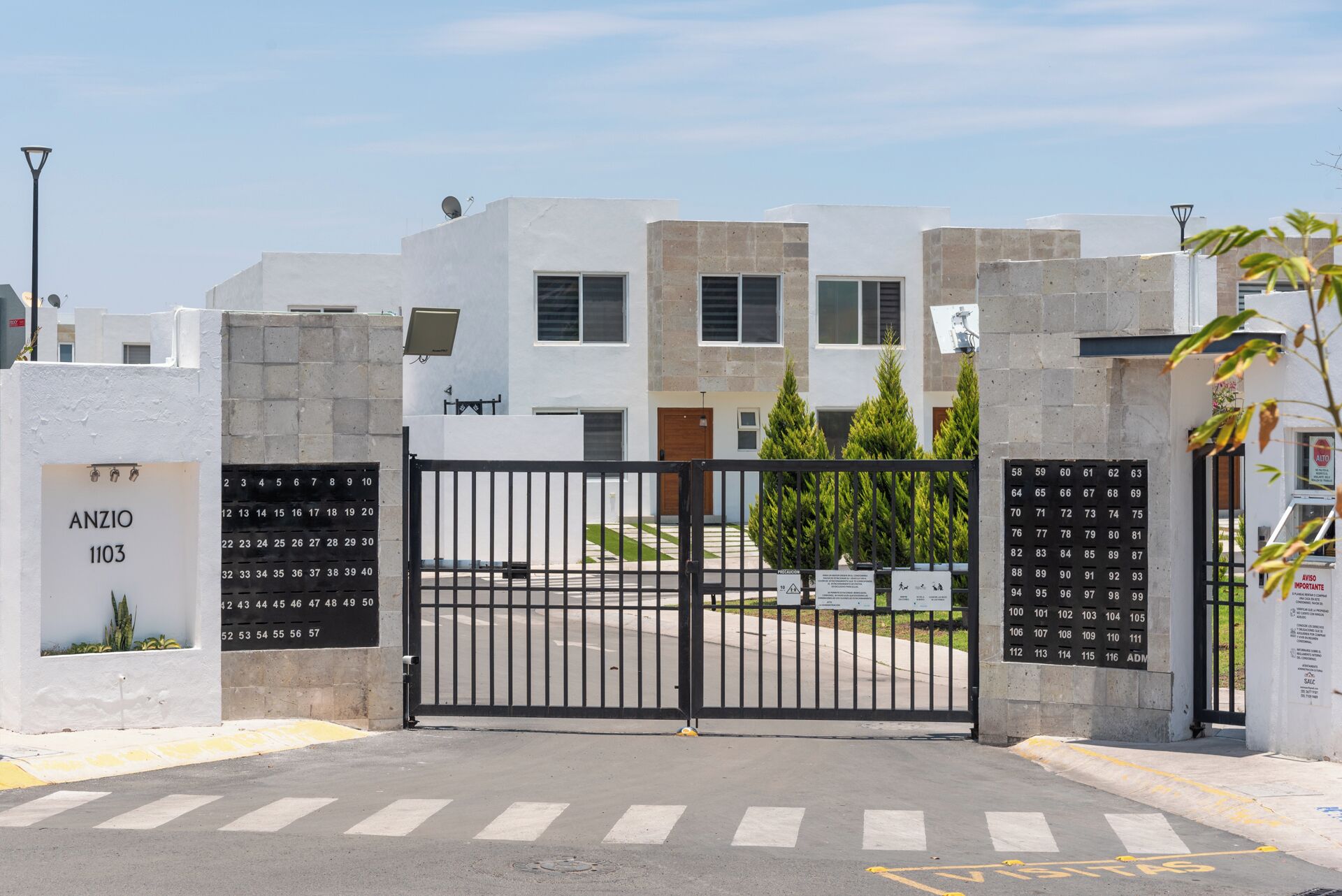 A gated residential complex with numbered apartment doors, a clear sky, and a pedestrian crossing.
