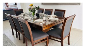 Mesa de jantar posta para seis com cadeiras, um vaso de flores e uma cozinha moderna ao fundo.