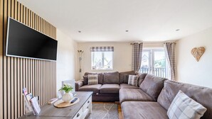 Un salon comprenant un canapé d’angle, un téléviseur à écran plat fixé sur un panneau en bois, une table basse avec un vase et des télécommandes, et une vue sur l’extérieur par une fenêtre.