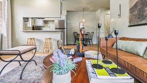 A modern living room with a sofa, a coffee table with books and decorative items, a dining area with chairs and a table, and a kitchen with cabinets and appliances.