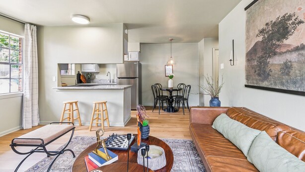 A modern living room with a sofa, a coffee table with books and a chess set, a dining area with chairs and a table, and a kitchen with a refrigerator and cabinets.
