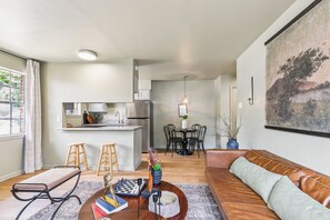 A modern living room with a sofa, a coffee table with books and a chess set, a dining area with chairs and a table, and a kitchen with a refrigerator and cabinets.