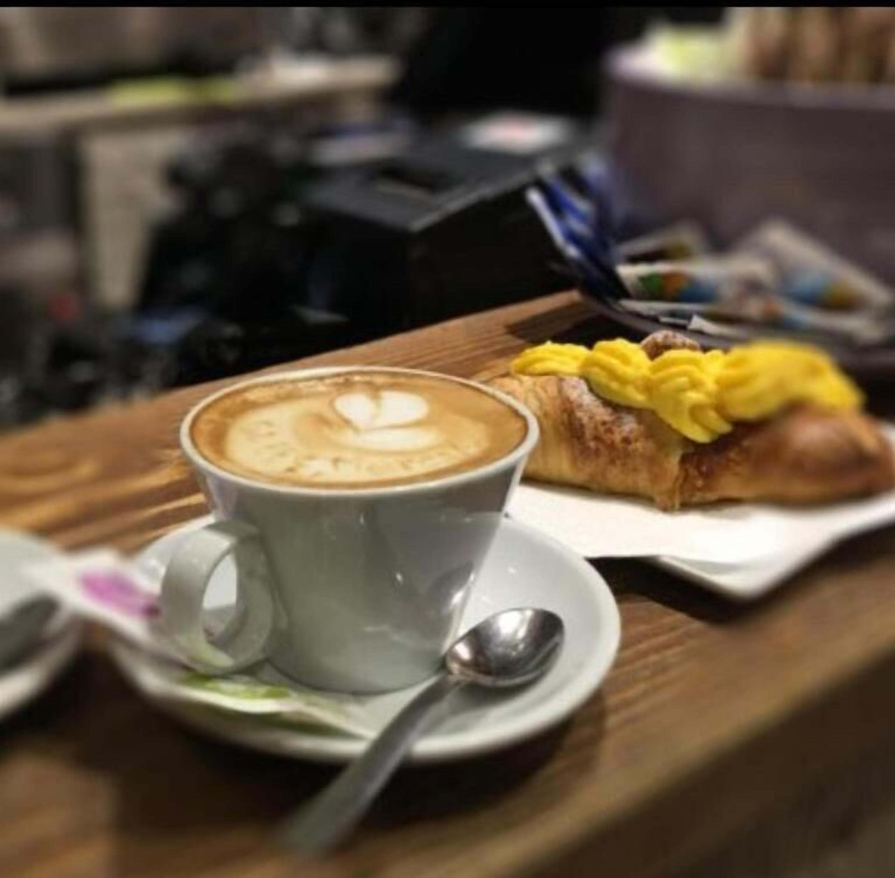 A cup of coffee with latte art on a saucer, a spoon, and a pastry with yellow topping on a plate.