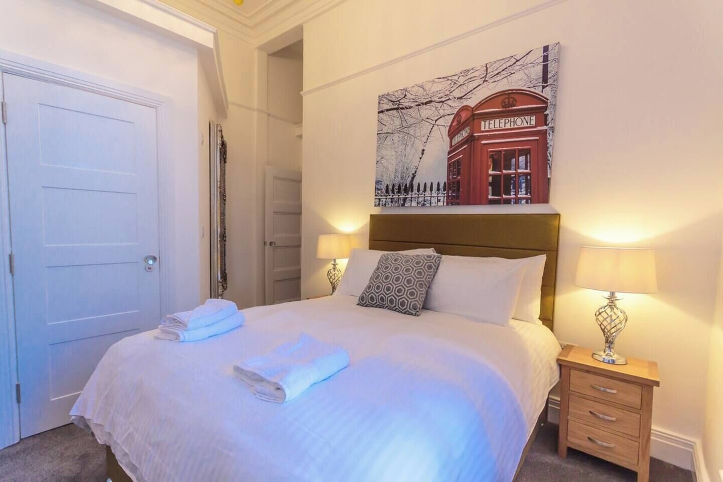 Une chambre d’hôtel avec un lit, deux lampes de chevet, une tête de lit et un tableau représentant une cabine téléphonique.