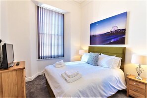 Ett hotellrum med en säng, ett skrivbord med en dator, ett fönster med persienner och en tavla på väggen som föreställer en stadsbild i nattljus.