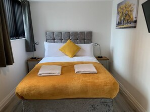 A neatly made bed with a headboard, two bedside tables, a window with curtains, and a painting on the wall.
