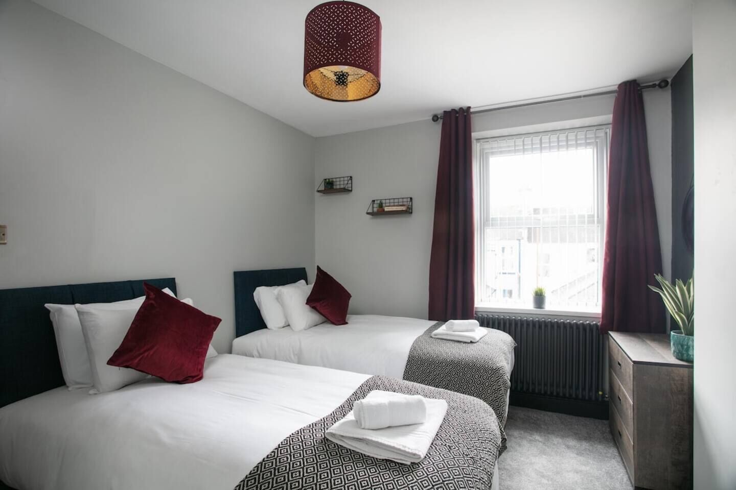 Une chambre d’hôtel avec deux lits, un radiateur, une fenêtre avec des rideaux et un luminaire suspendu.