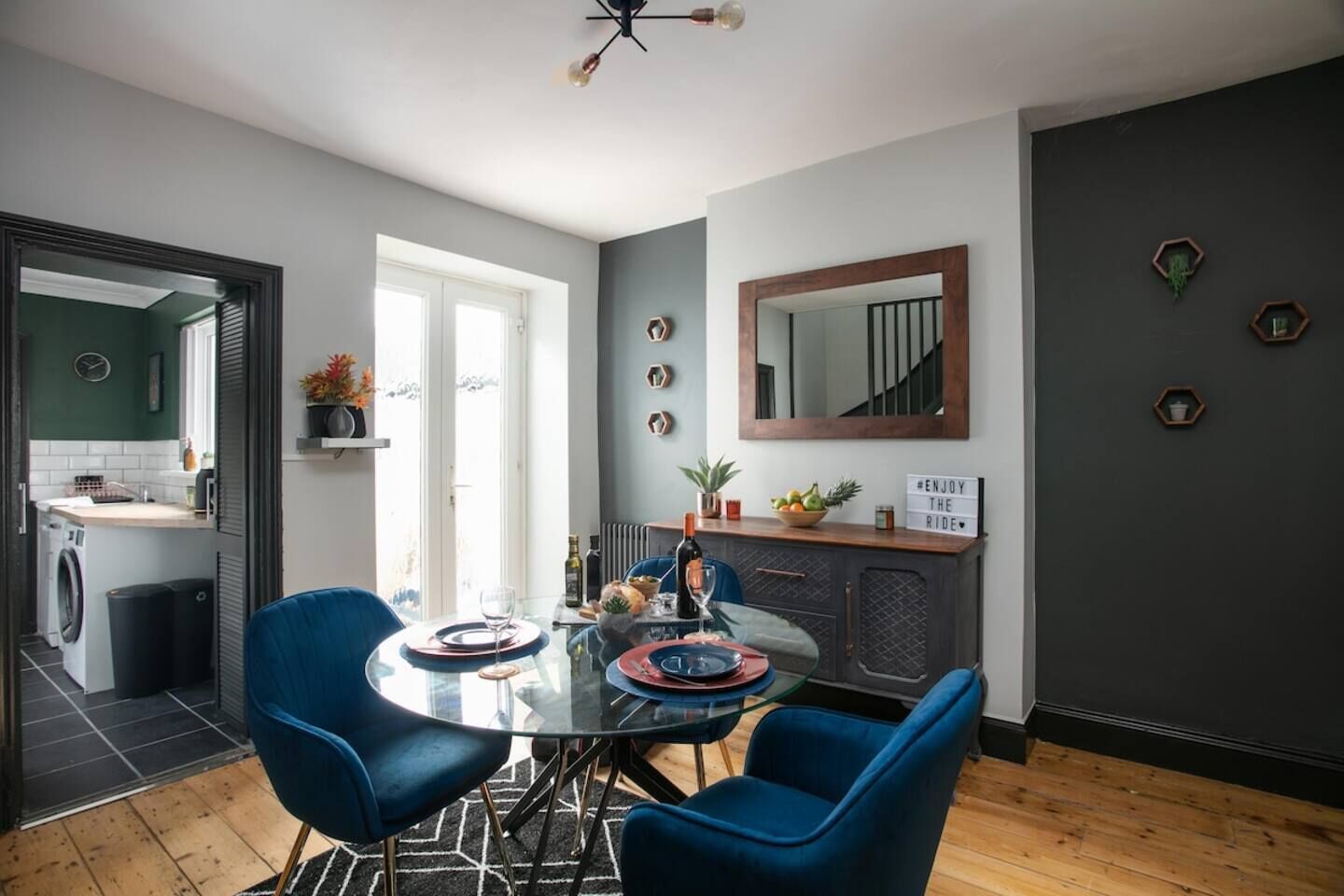 Een moderne keuken met een eethoek, met een glazen tafel, blauwe stoelen en een donkergrijze wand met decoratieve honingraatspiegels.