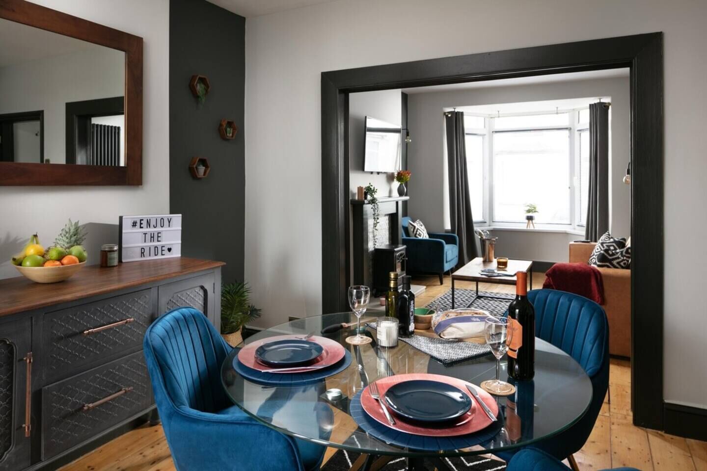 A dining area with a glass table set for four, blue chairs, and a dark wooden cabinet with a bowl of fruit on top.