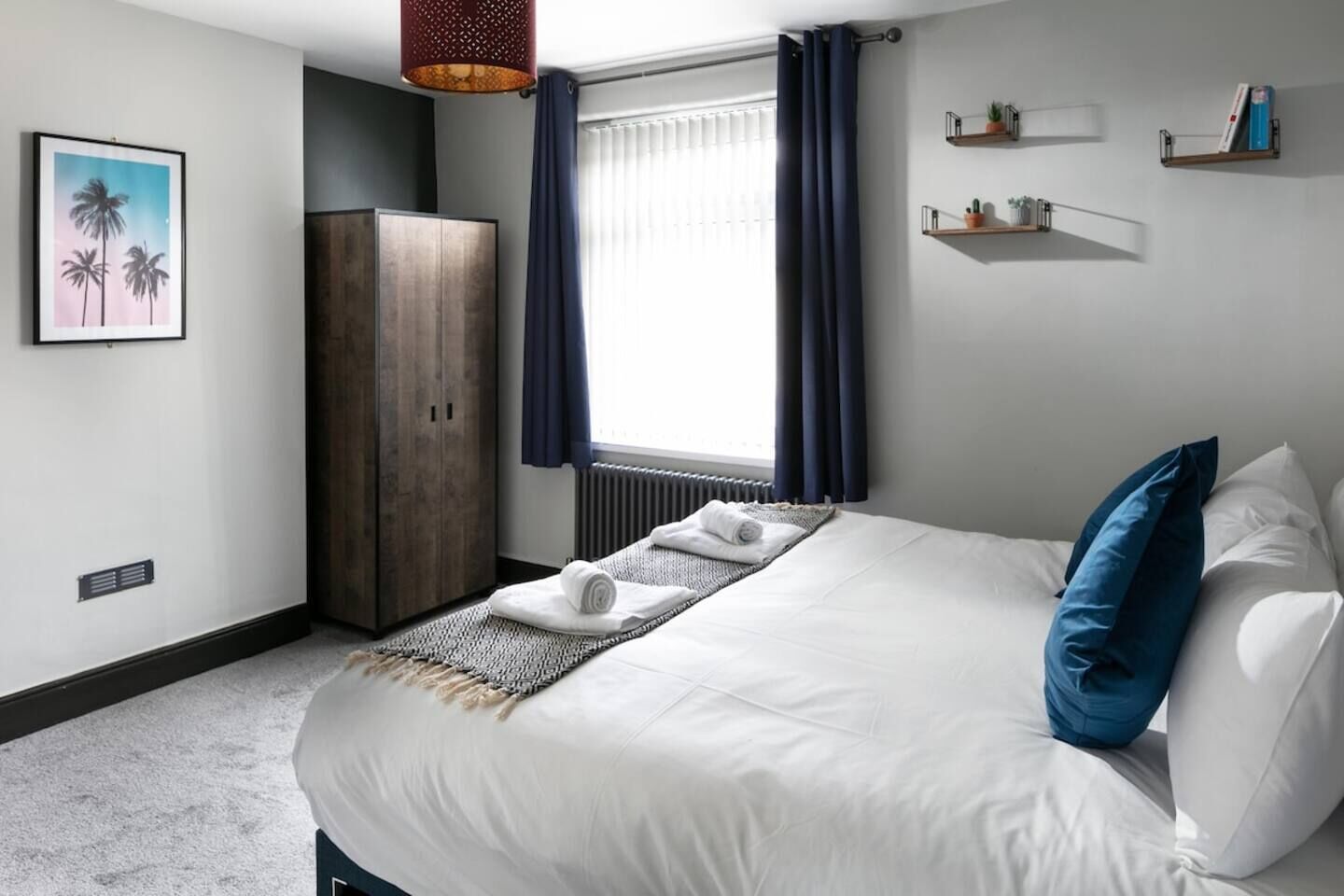 A hotel room with a bed, a wardrobe, a radiator, a window with curtains, and wall-mounted shelves with plants.