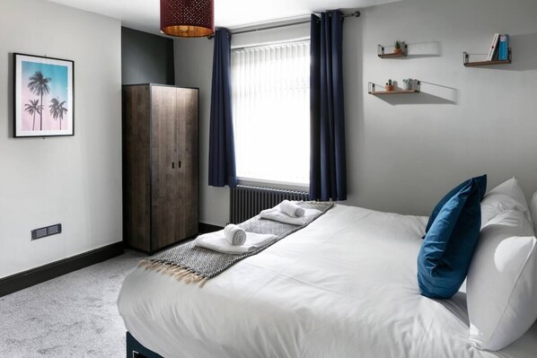 A hotel room with a bed, a wardrobe, a radiator, a window with curtains, and wall-mounted shelves with plants.