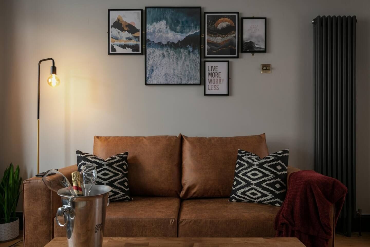 A living room with a brown sofa, patterned cushions, a floor lamp, a radiator, and framed artwork on the wall.