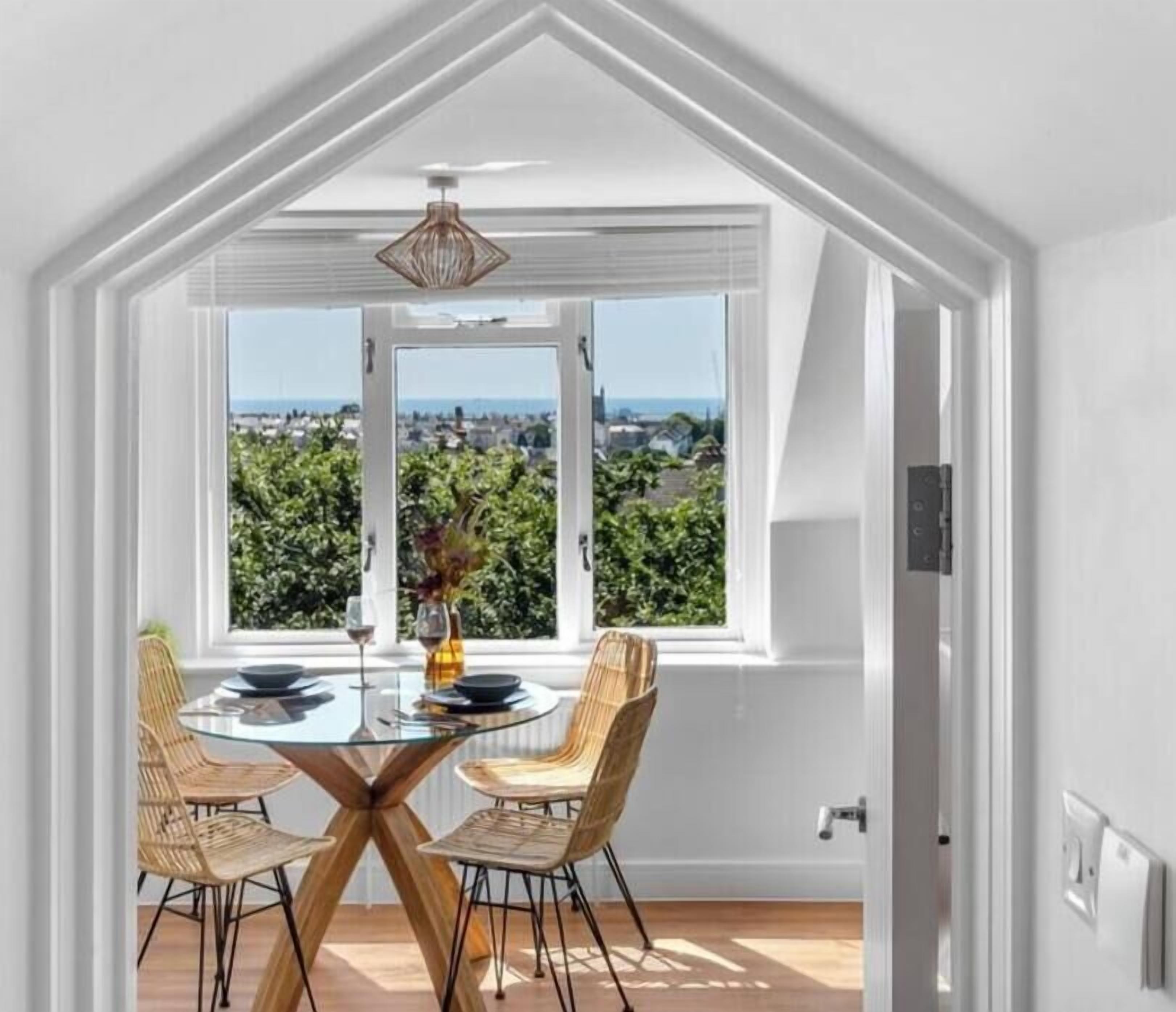 A dining area with a glass table and wicker chairs, a view of greenery and buildings through large windows, and a white ceiling with a hanging light fixture.