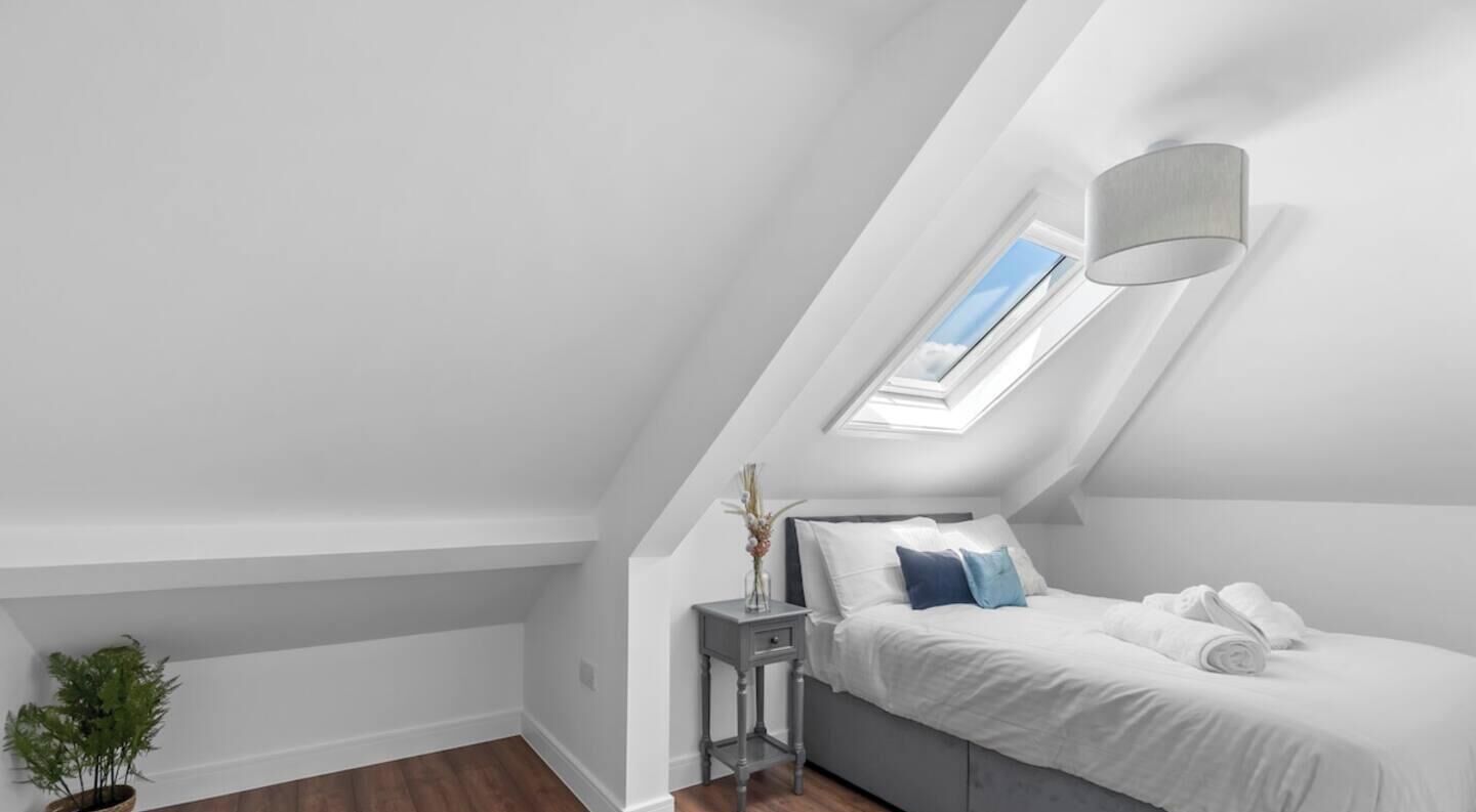 Un dormitorio con tragaluz, una mesita de noche con un florero, y una cama con ropa de cama blanca.