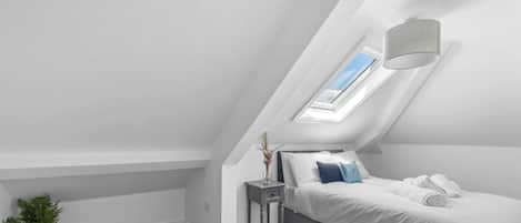 A bedroom with a skylight, a bedside table with a vase, and a bed with white linens.