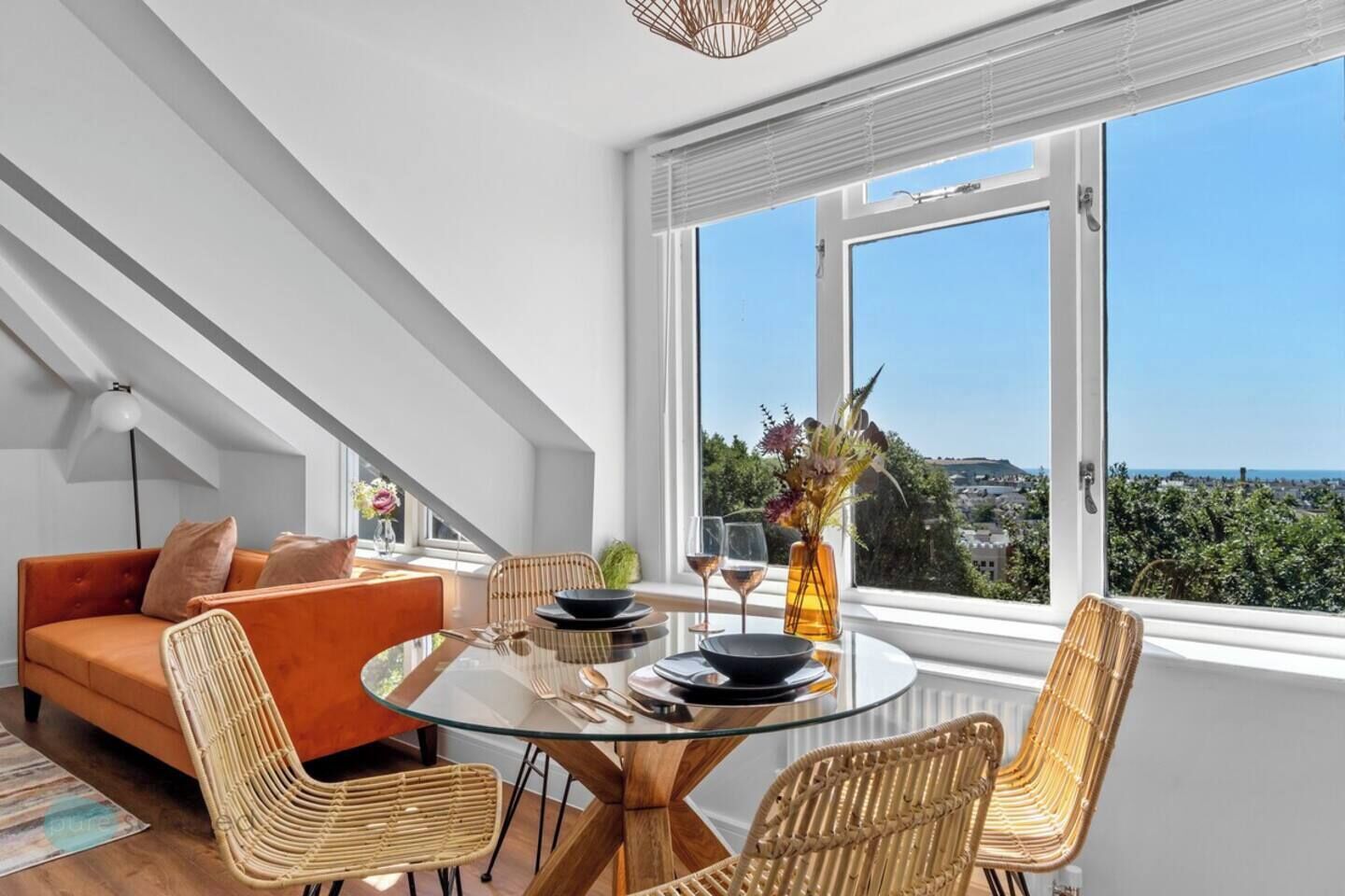 Un espace repas moderne avec une table en verre, des chaises en osier et une vue sur la ville grâce à de grandes fenêtres.
