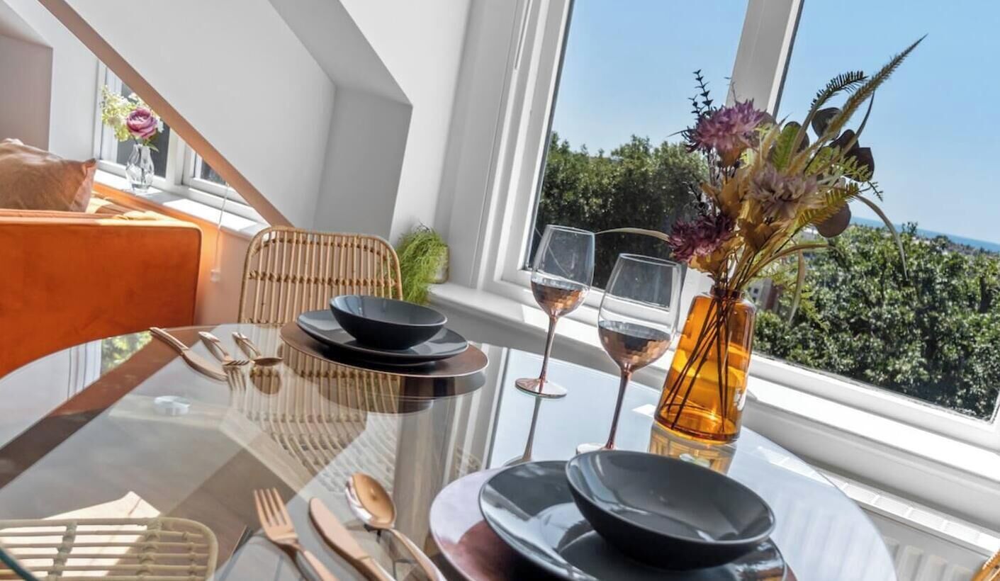 A dining table with black plates, cutlery, and two wine glasses, set against a window with a view of greenery and the sea.