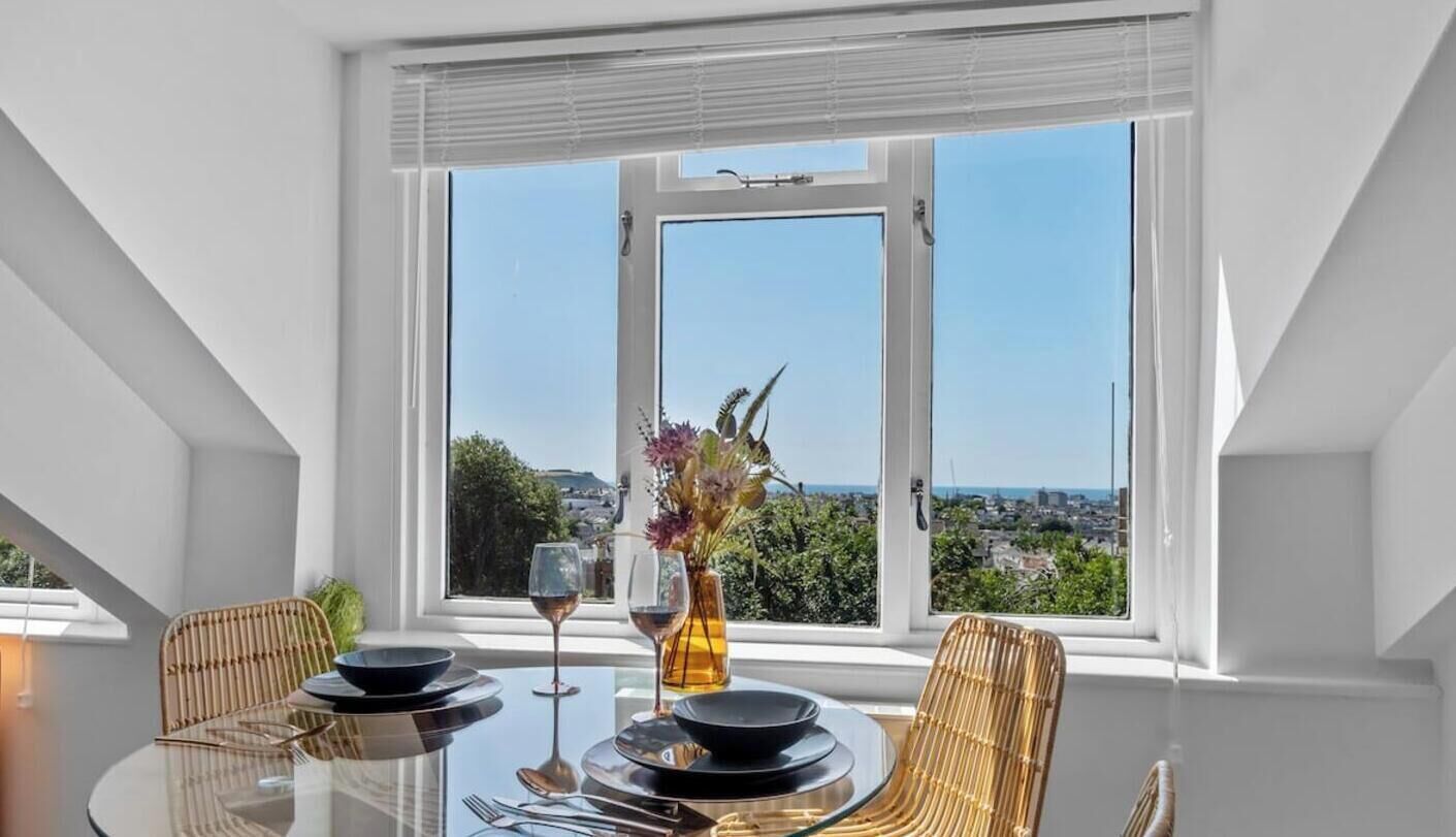 A dining area with a table set for two, a window with a view of a cityscape, and a vase with flowers on the table.
