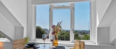 A dining area with a table set for two, a window with a view of a cityscape, and a vase with flowers on the table.