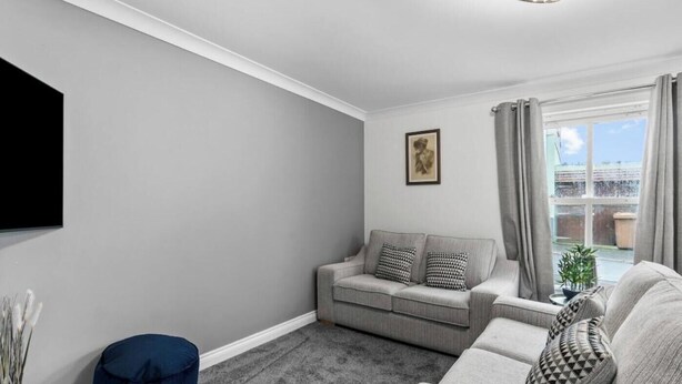 Una sala de estar con un sofá gris, un reposapiés azul y un televisor de pantalla plana montado en la pared.