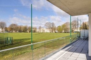 A view from a balcony overlooking a sports field with a netted fence, a paved walkway, and a building in the background. - Luxury 3 bed apartment Mount Wise Plymouth parking (Plymouth)
