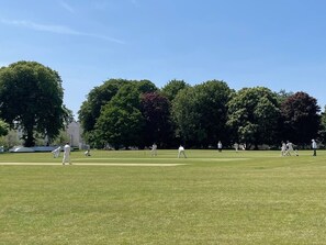 An outdoor cricket field with players in white attire, surrounded by trees and a clear sky. - Luxury 3 bed apartment Mount Wise Plymouth parking (Plymouth)