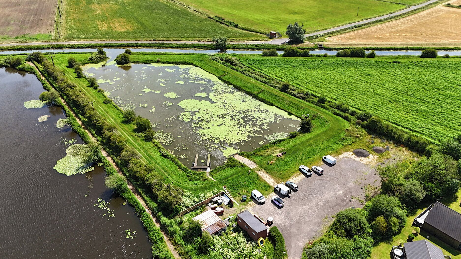 Luchtbeeld van een landgoed met een vijver, groene velden en geparkeerde voertuigen.