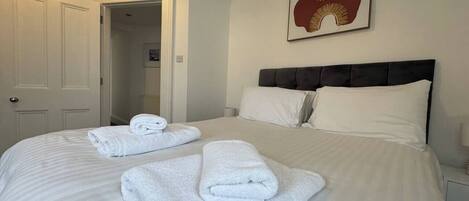A hotel room with a large bed, two rolled towels, a headboard, a nightstand, and a framed artwork on the wall.