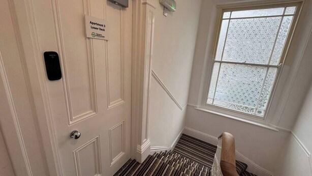 Entrada de hotel con un cartel que dice 'Apartment 3 Lower Deck', una puerta con ranura para tarjeta de acceso, un ventanal con vidrio emplomado y una alfombra a rayas.