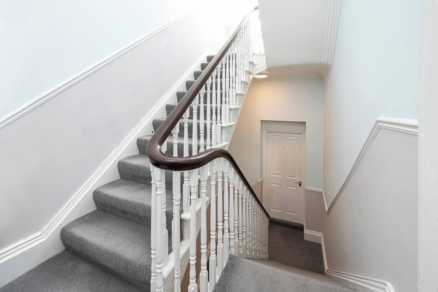 Un escalier avec des balustrades blanches et une main courante sombre, menant à un palier avec une porte fermée.